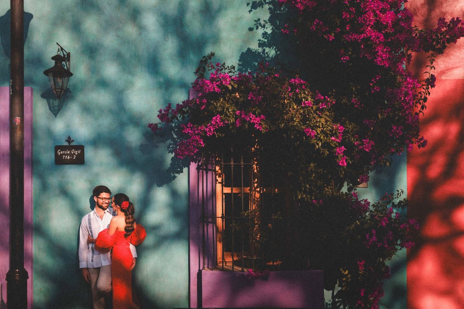 Couple walking through a garden path with vibrant pink bougainvillea flowers and glowing sunset lighting.