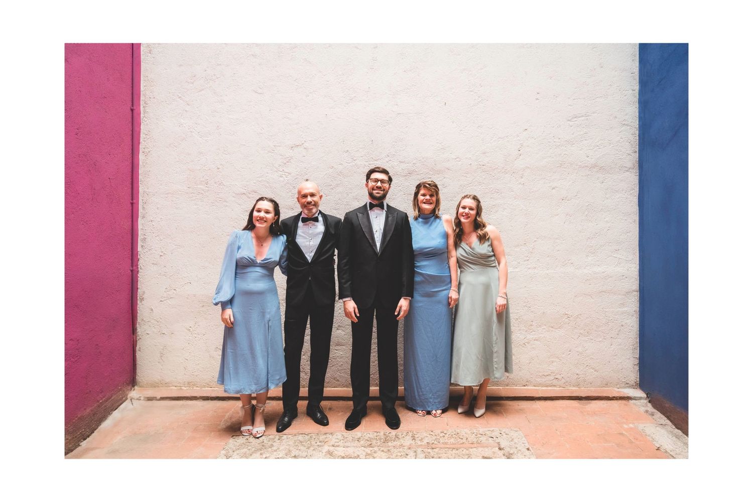 Wedding party group photo against white wall with pink and blue accent colors.