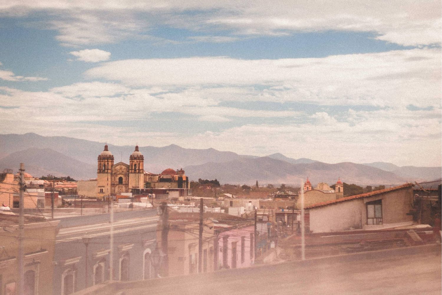 Panoramic desert landscape with adobe buildings and mountains under a cloudy sky at sunset.