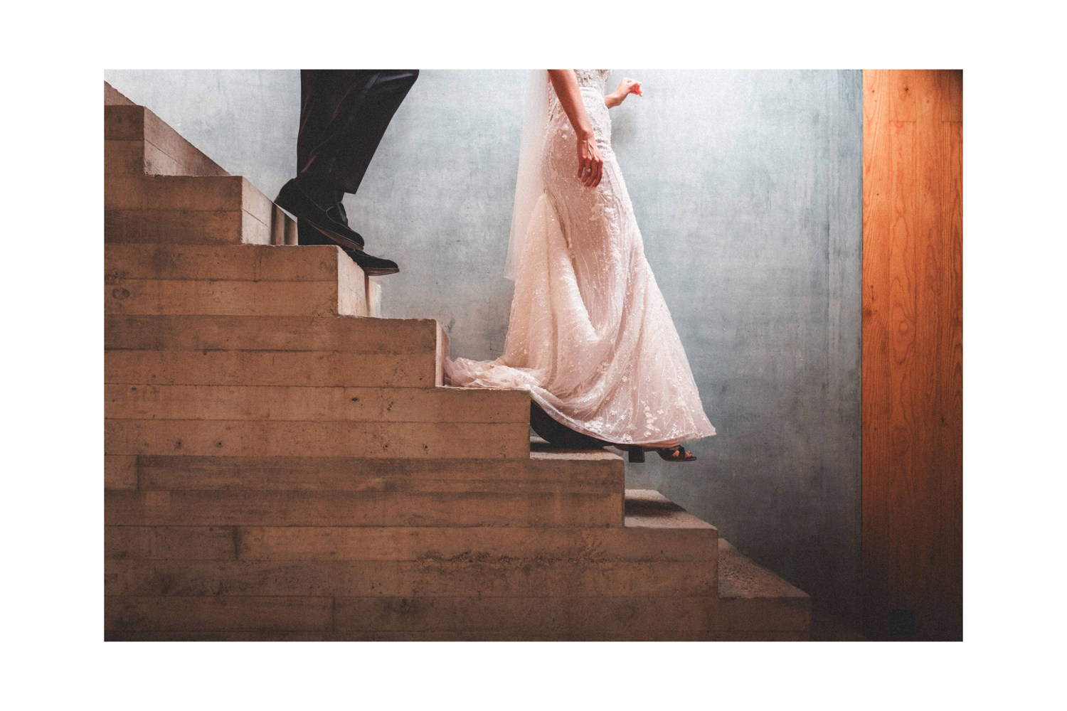 A person in a flowing pink dress and black heels ascends wooden stairs against a concrete wall.