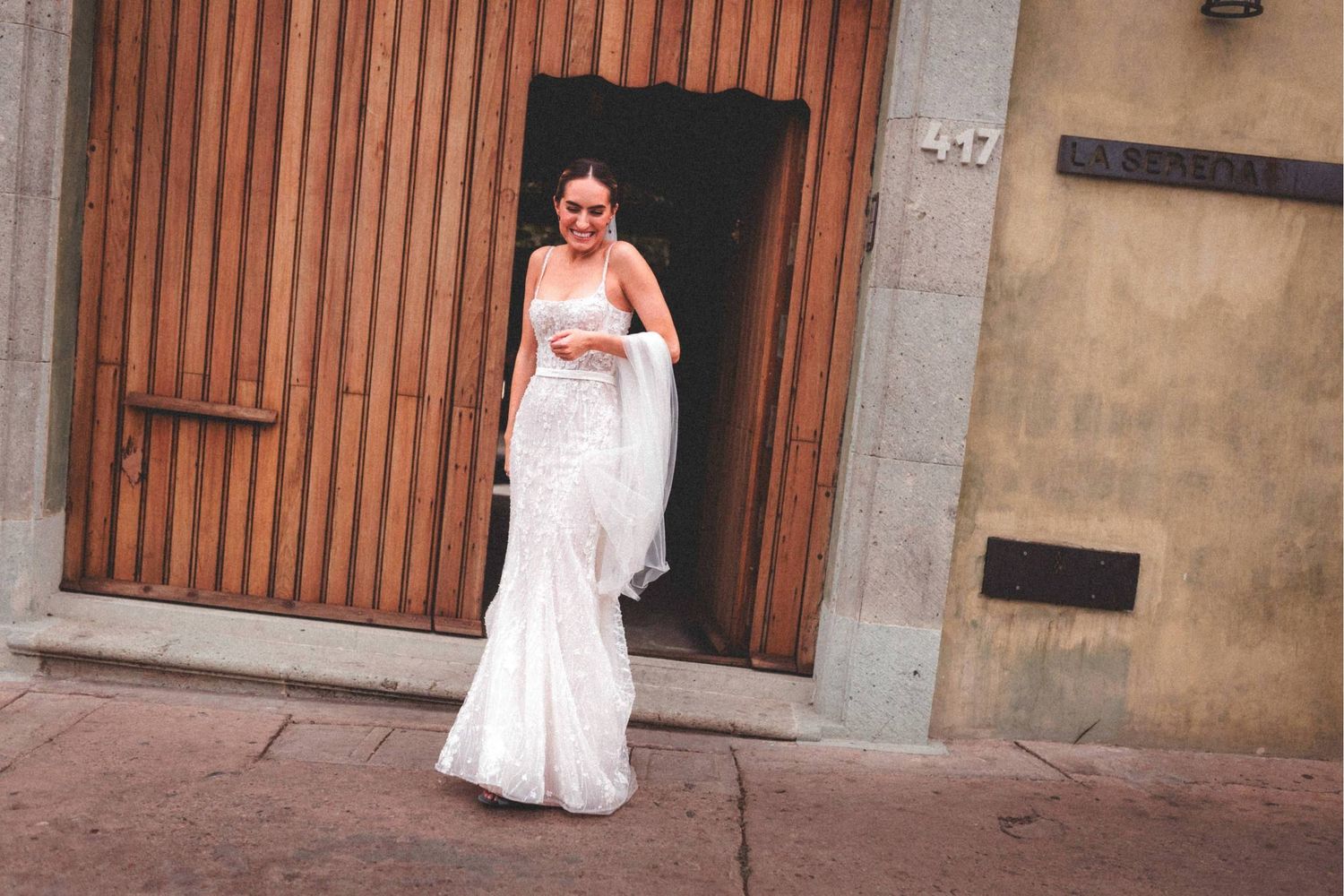 A person in a white wedding dress poses against a rustic stone wall with wooden doors in the background.