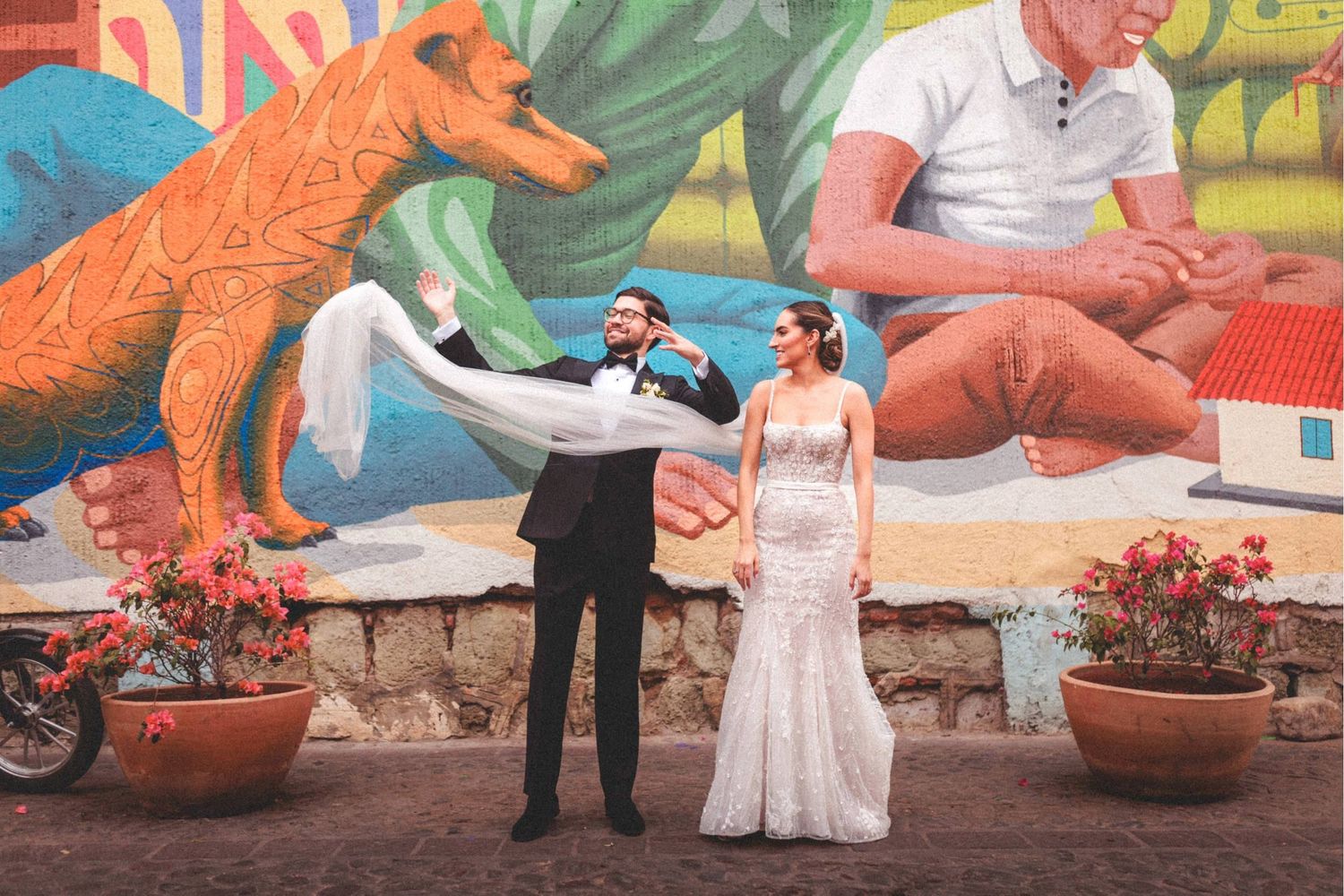 A couple poses for wedding photos in front of a vibrant orange dinosaur statue at a unique outdoor venue.