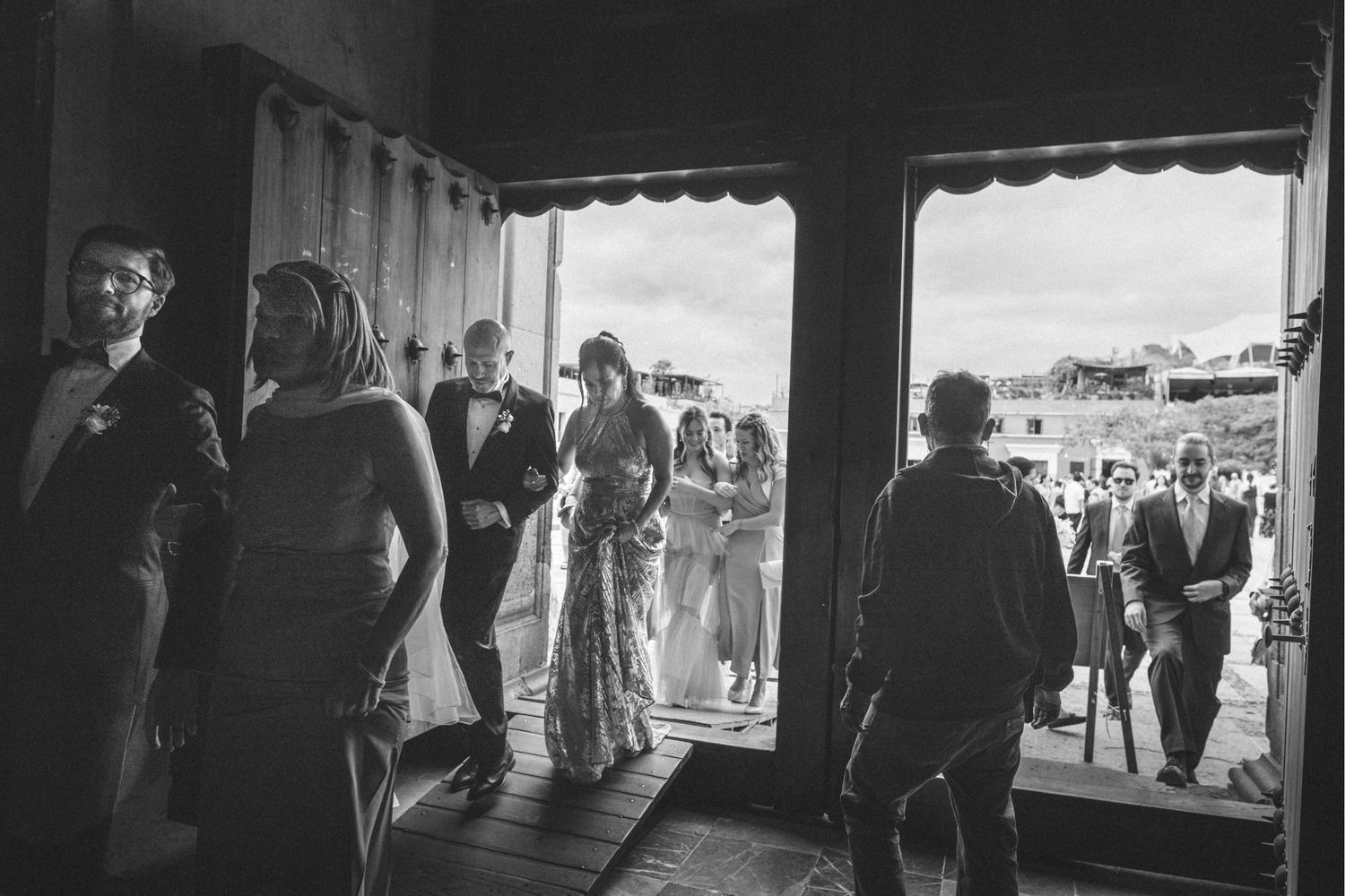Silhouettes of wedding guests mingle in an elegant reception venue with large windows overlooking a scenic view.