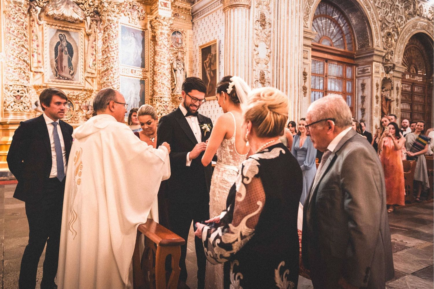Wedding guests mingle and socialize during a reception in an ornate baroque-style ballroom with gilded decor.