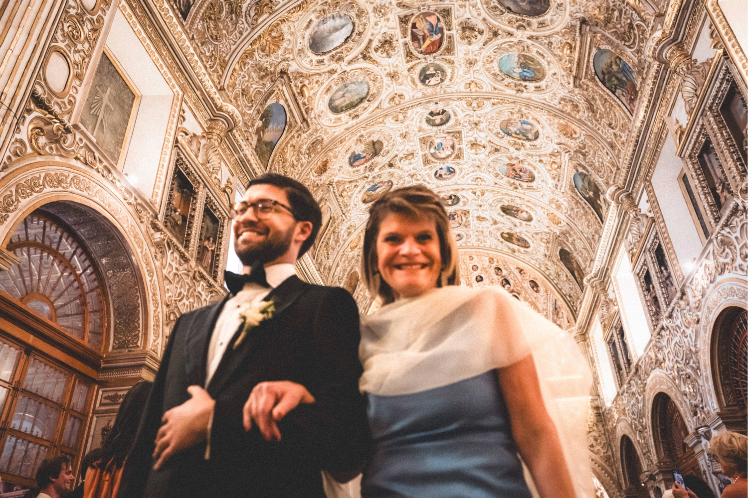 Wedding party members laughing together in an ornate baroque church interior with decorated ceiling and columns.