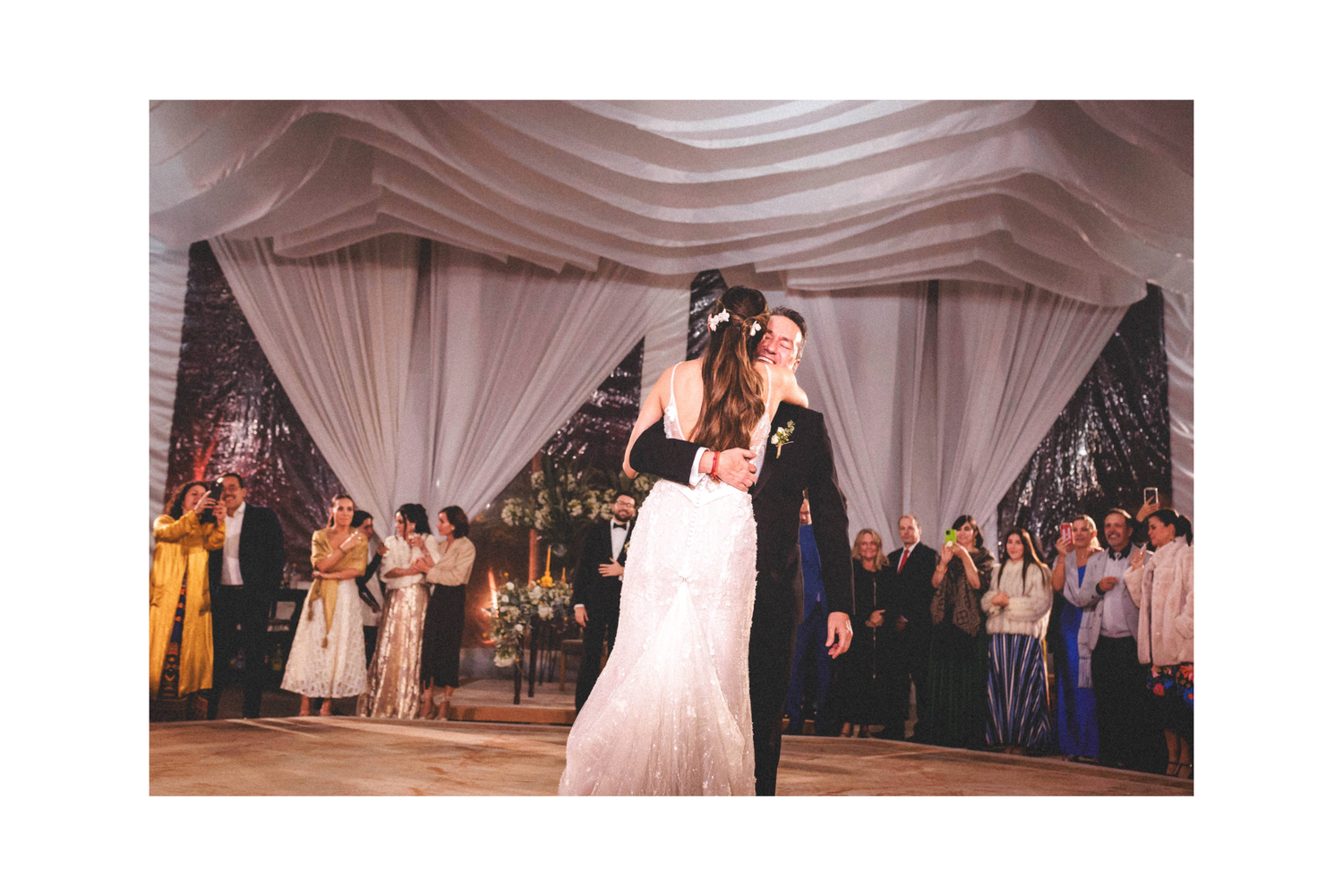 Newlyweds share their first dance under elegant white draped fabric at an indoor wedding reception with guests watching.
