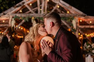 A couple shares a romantic kiss during their first dance at a rustic barn wedding reception with twinkling lights.