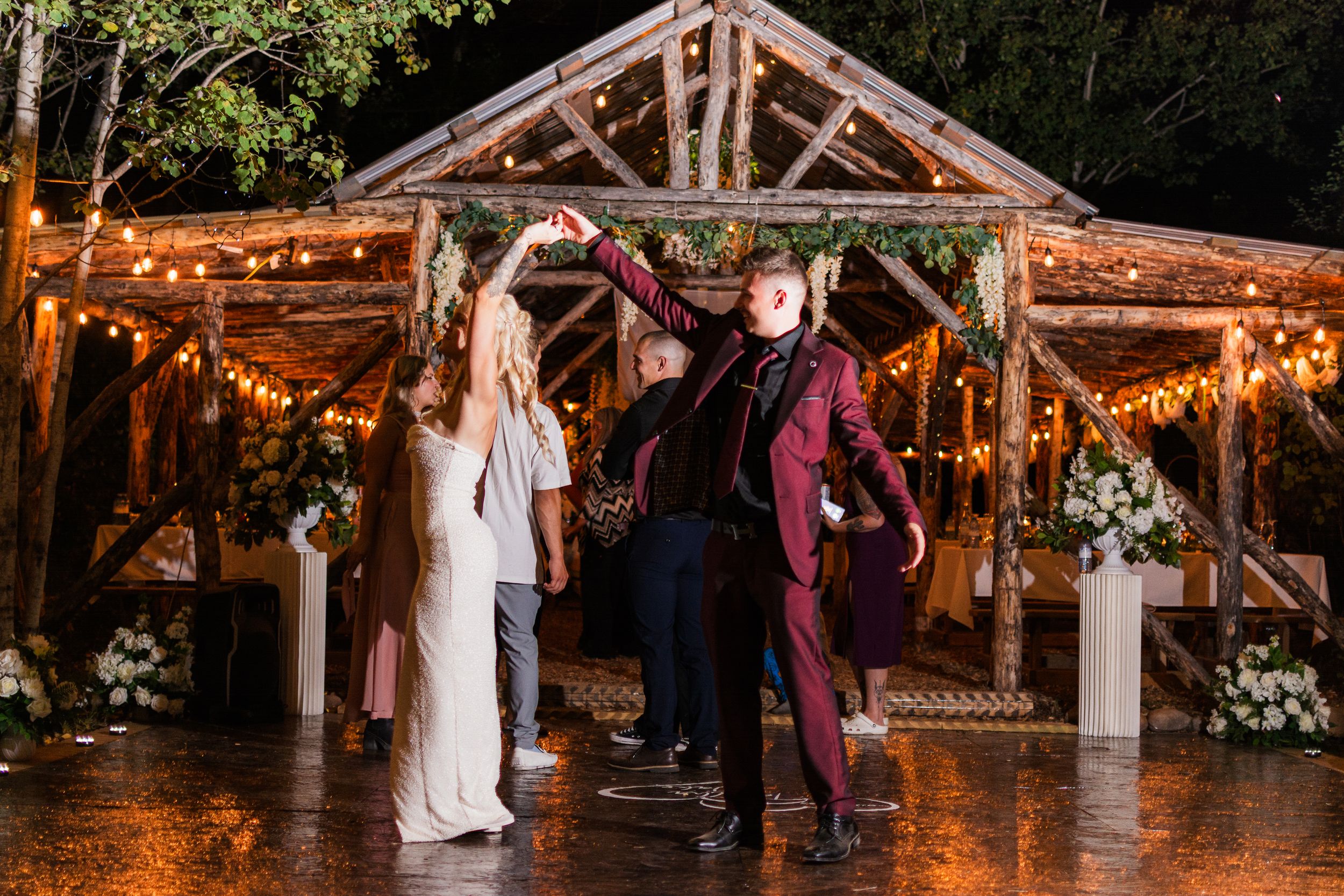 A romantic couple dances beneath string lights at a rustic outdoor wedding reception pavilion.