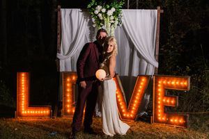 Romantic outdoor wedding scene with glowing LOVE marquee letters and white curtains against dark foliage at night.