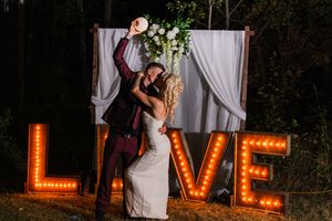 A romantic outdoor wedding scene with glowing LOVE marquee letters and white curtains in the background.