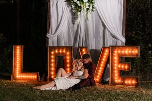Large illuminated LOVE marquee letters displayed against white drapes and greenery at an evening event.