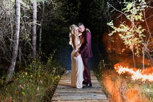 A couple shares an intimate moment on a wooden walkway in the woods with glowing lights in the background.