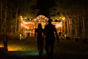 String lights illuminate a wooded outdoor wedding venue at night with guests mingling beneath the trees.