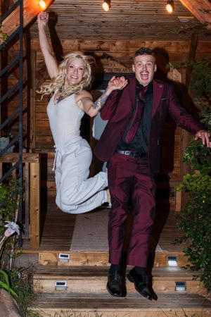 Energetic couple jumping together on wooden steps decorated with string lights.