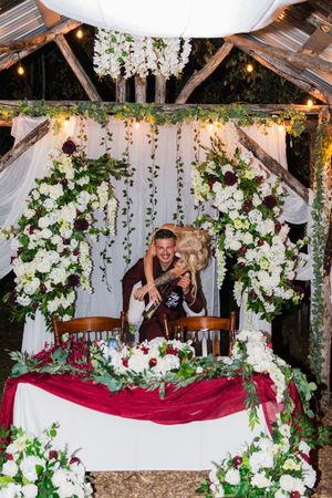 A couple celebrates with champagne under a floral-decorated wedding arch with white draping and burgundy accents.