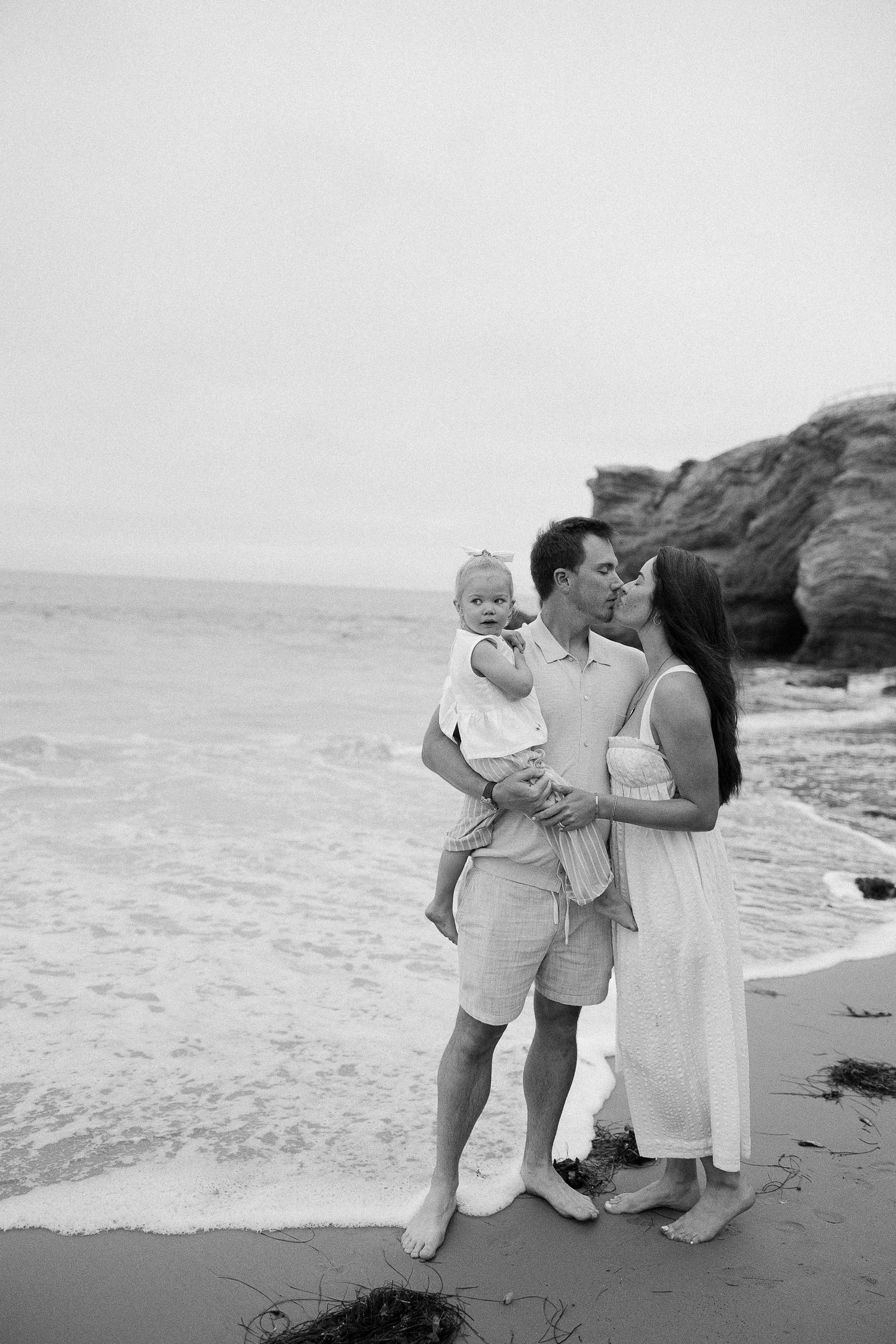 A family in light colored clothing embraces on a rocky beach as ocean waves crash behind them in black and white.