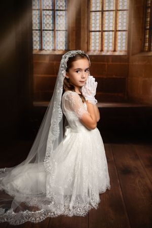 A young girl in a white lace communion dress and veil stands in front of ornate church windows on hardwood floors.