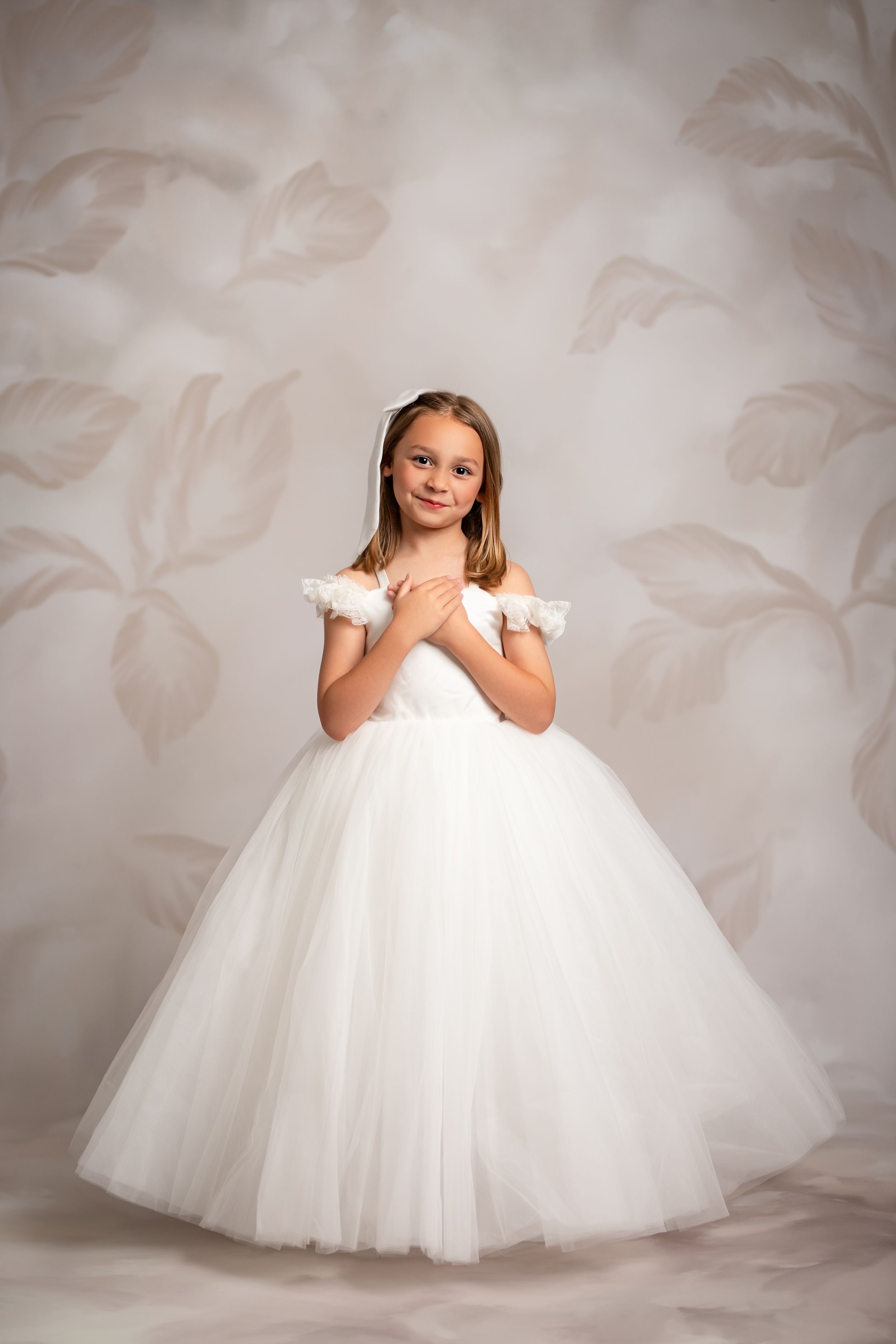 Young child in an elegant white ball gown with tulle skirt poses in front of a floral backdrop.