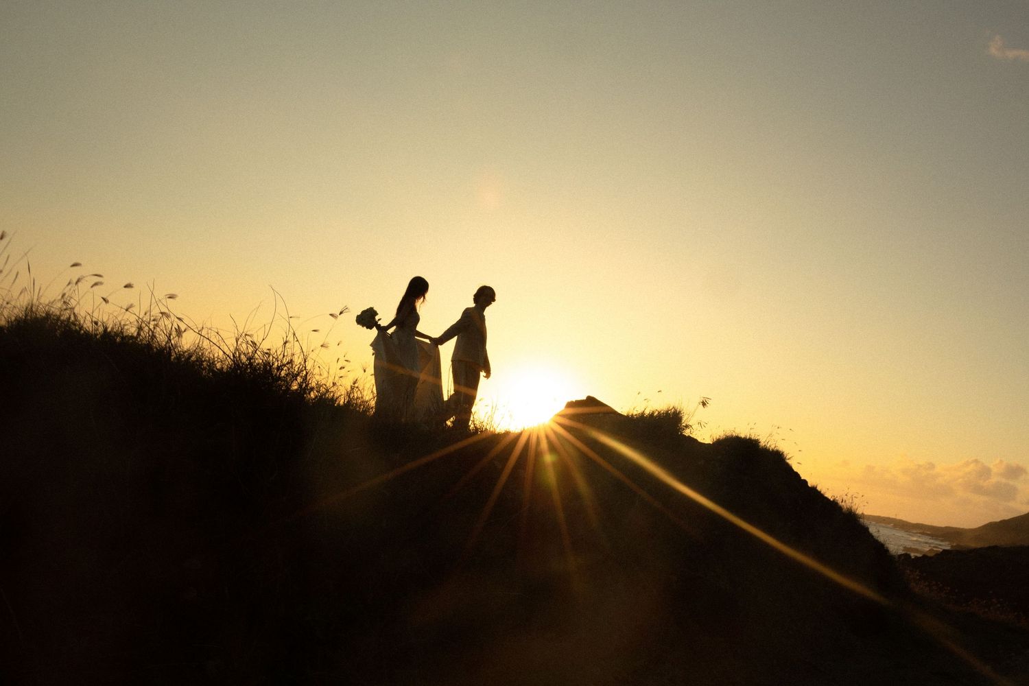 Silhouetted couple in wedding attire stands on rocky shore against dramatic golden sunset over ocean.