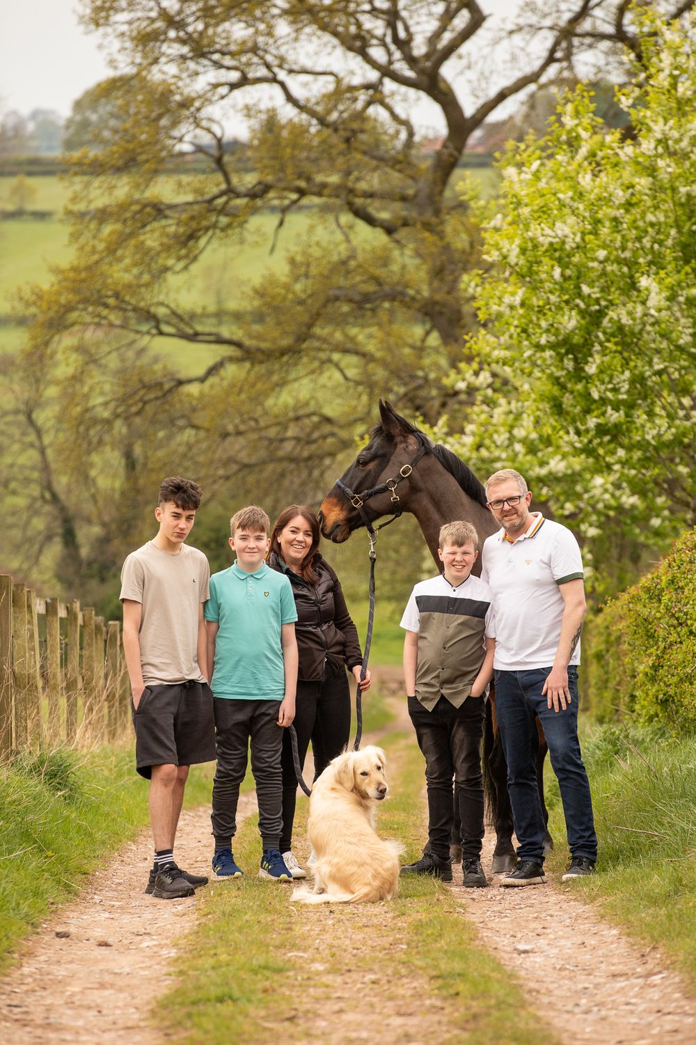 A group walks along a rural path with their black horse and golden retriever dog on a sunny day.