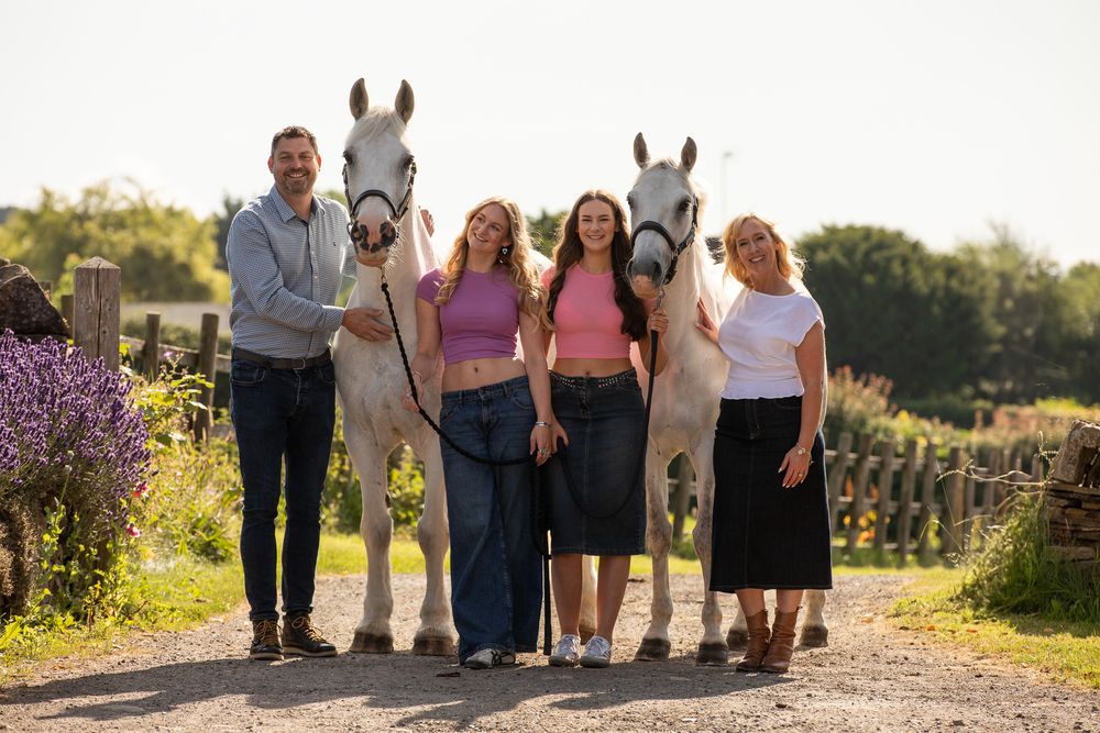 A group of friends pose with two beautiful white horses on a scenic country path at sunset.