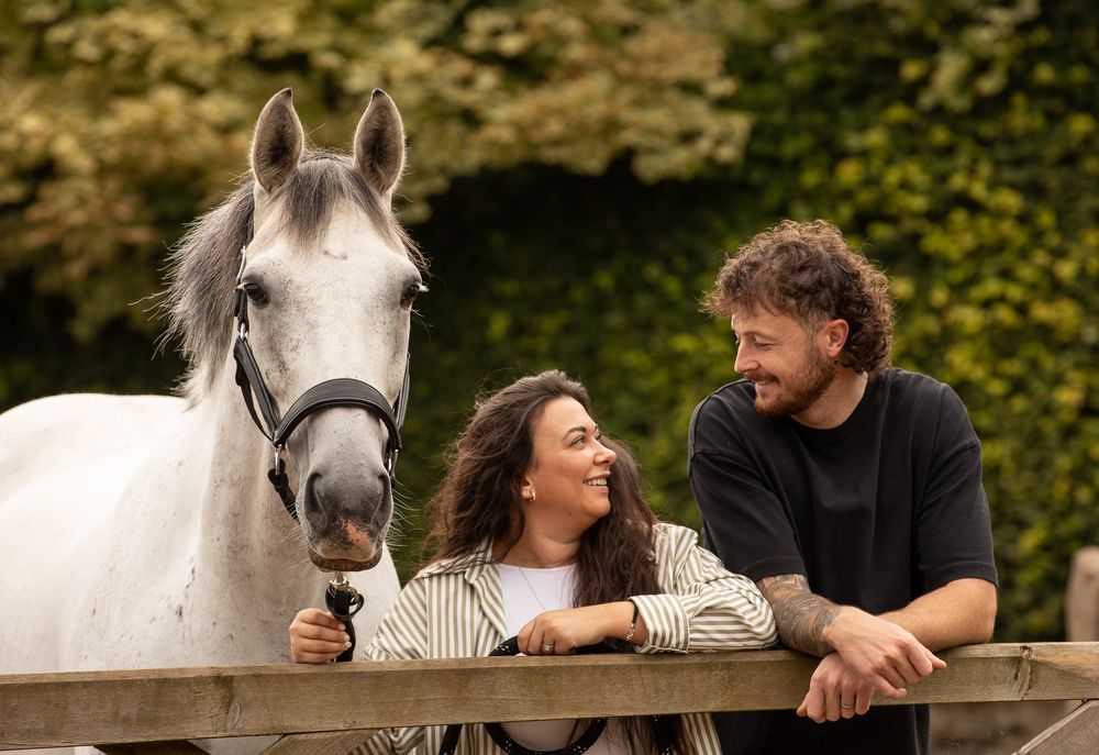 A beautiful white horse stands beside a wooden fence as a couple shares a warm moment together outdoors.