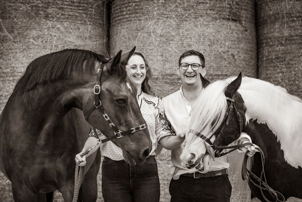 A couple poses with two horses, one white and one brown, against a brick wall in a black and white photograph.