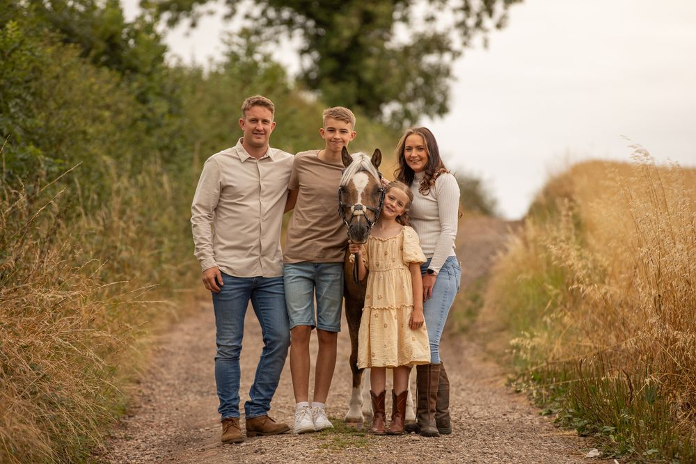 A family of four standing together on a dirt path surrounded by tall grass and golden fields on a warm afternoon.