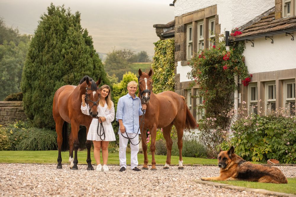 A group stands with two brown horses in front of a charming country house while a dog rests nearby.