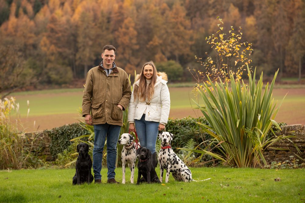 Couple standing on grass with three dogs including a black Lab, Border Collie and Dalmatian in autumn setting.