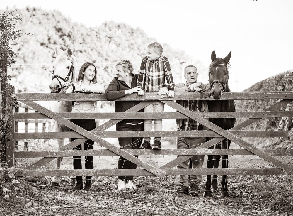 A vintage black and white photograph shows several people gathered at a wooden farm fence alongside two horses.