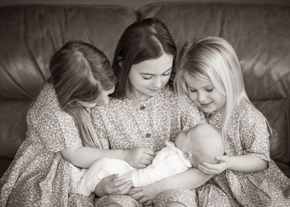 Three young girls in matching floral dresses sit together on a leather couch gazing at a baby in white clothing.