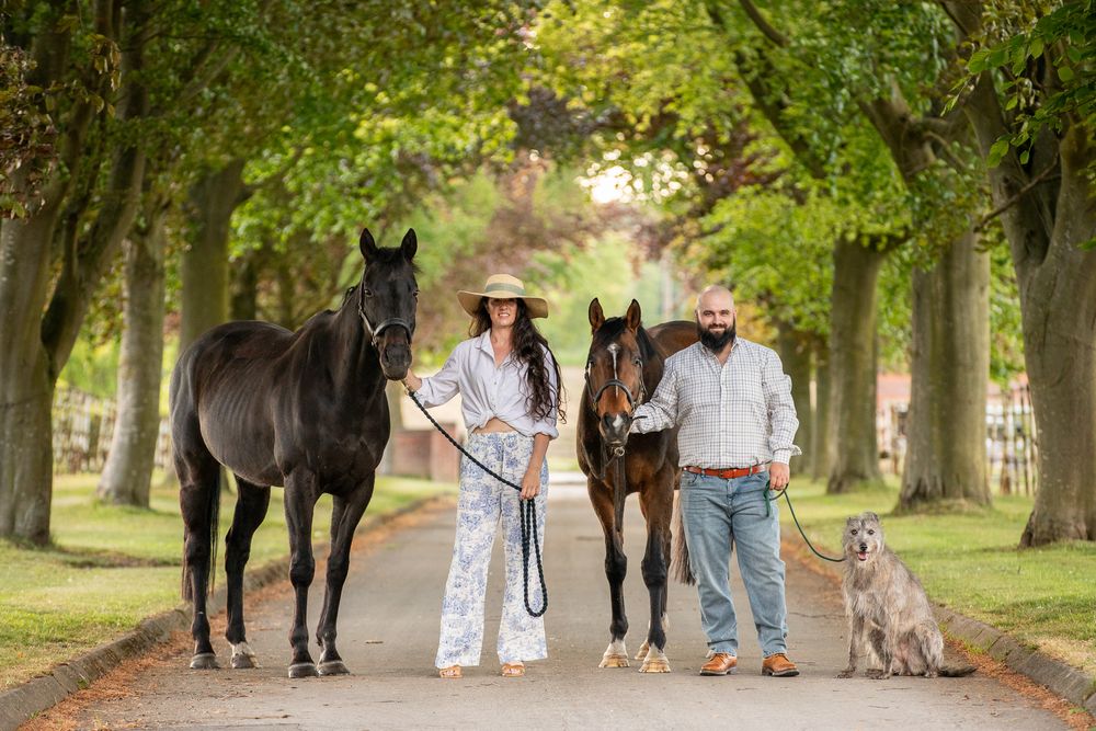 Two people stand with their horses and dog on a tree-lined path during a sunny afternoon.
