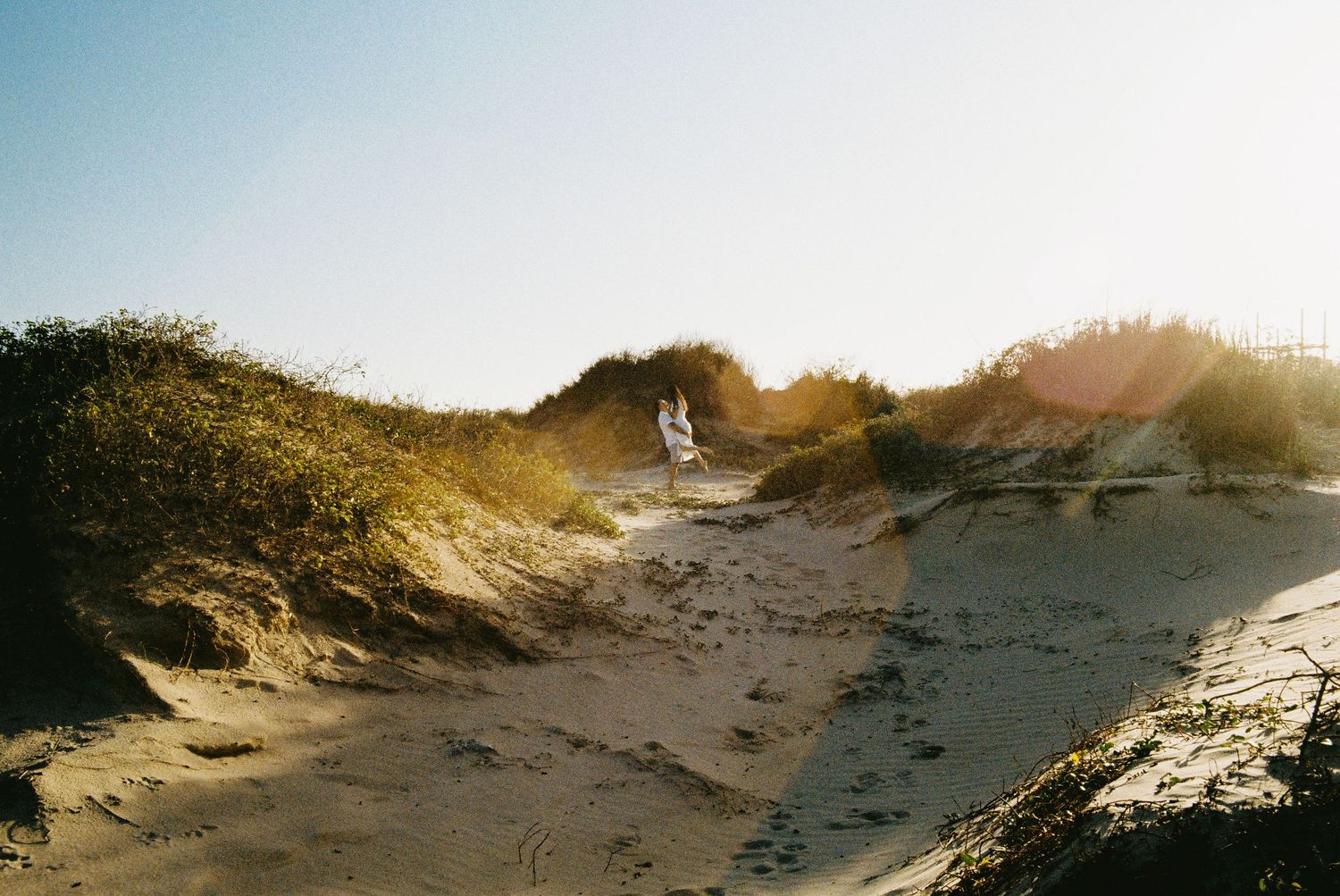 Woman in white dress walking along sandy beach dunes on a clear sunny day, shot on Kodak Portra 400. SPI Photoshoot.