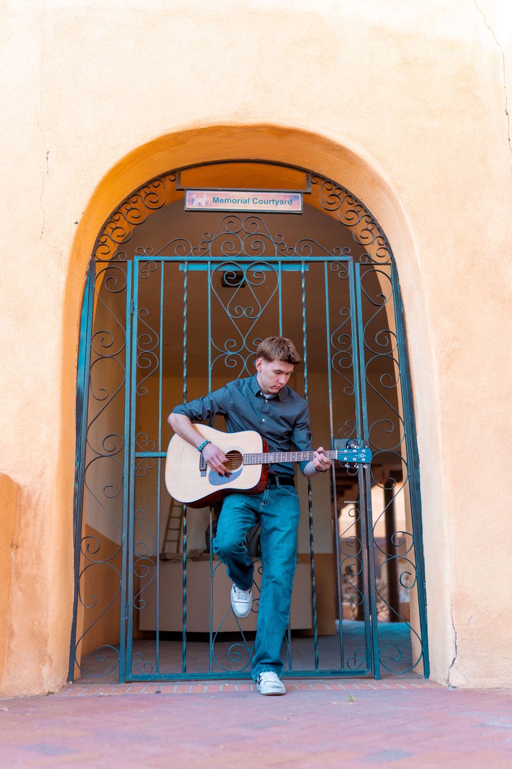 Musician playing acoustic guitar while leaning against decorative iron gate in arched doorway.