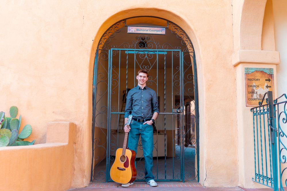 Musician in blue shirt stands in arched doorway with acoustic guitar against peach colored wall.