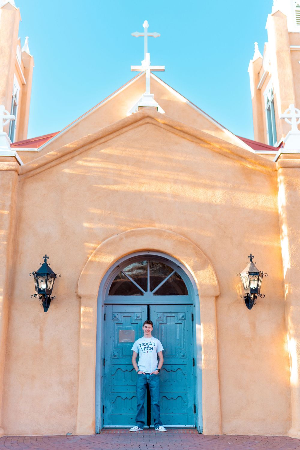 Person standing in front of turquoise church door with cross and lanterns on peach building.
