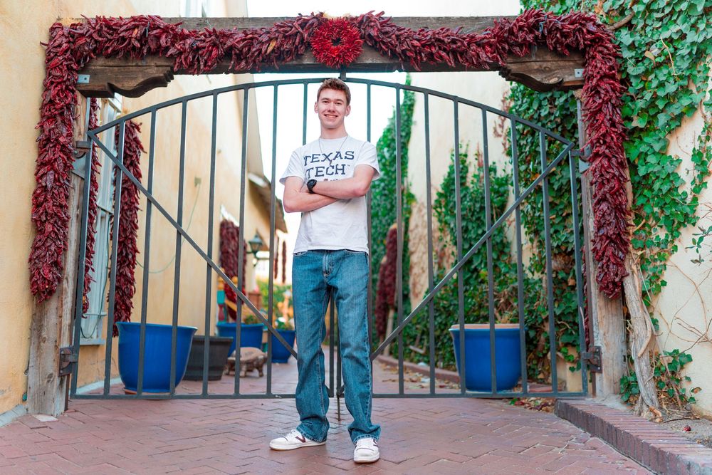 Person in casual wear standing under decorative archway with red vines and blue planters.