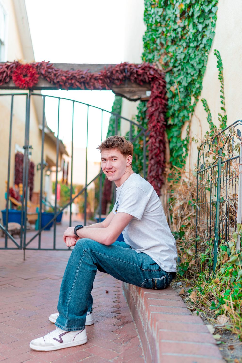 Person in white t-shirt sitting on ledge with ivy covered wall and metal gate behind.