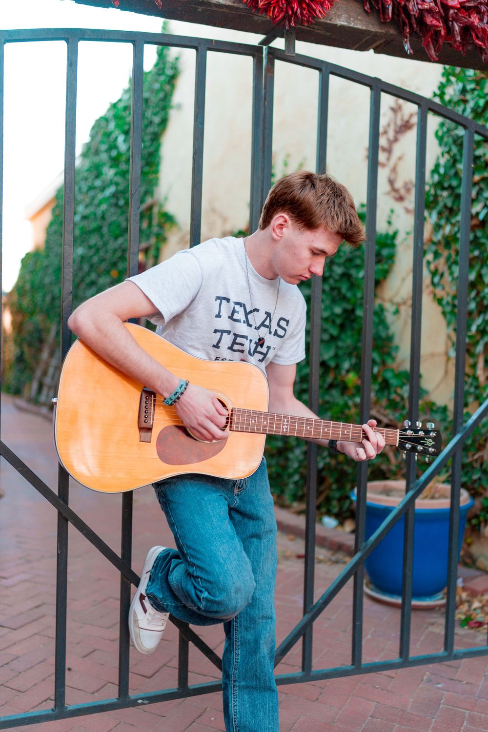 Musician in Texas Tech shirt playing acoustic guitar against metal gate and ivy wall.