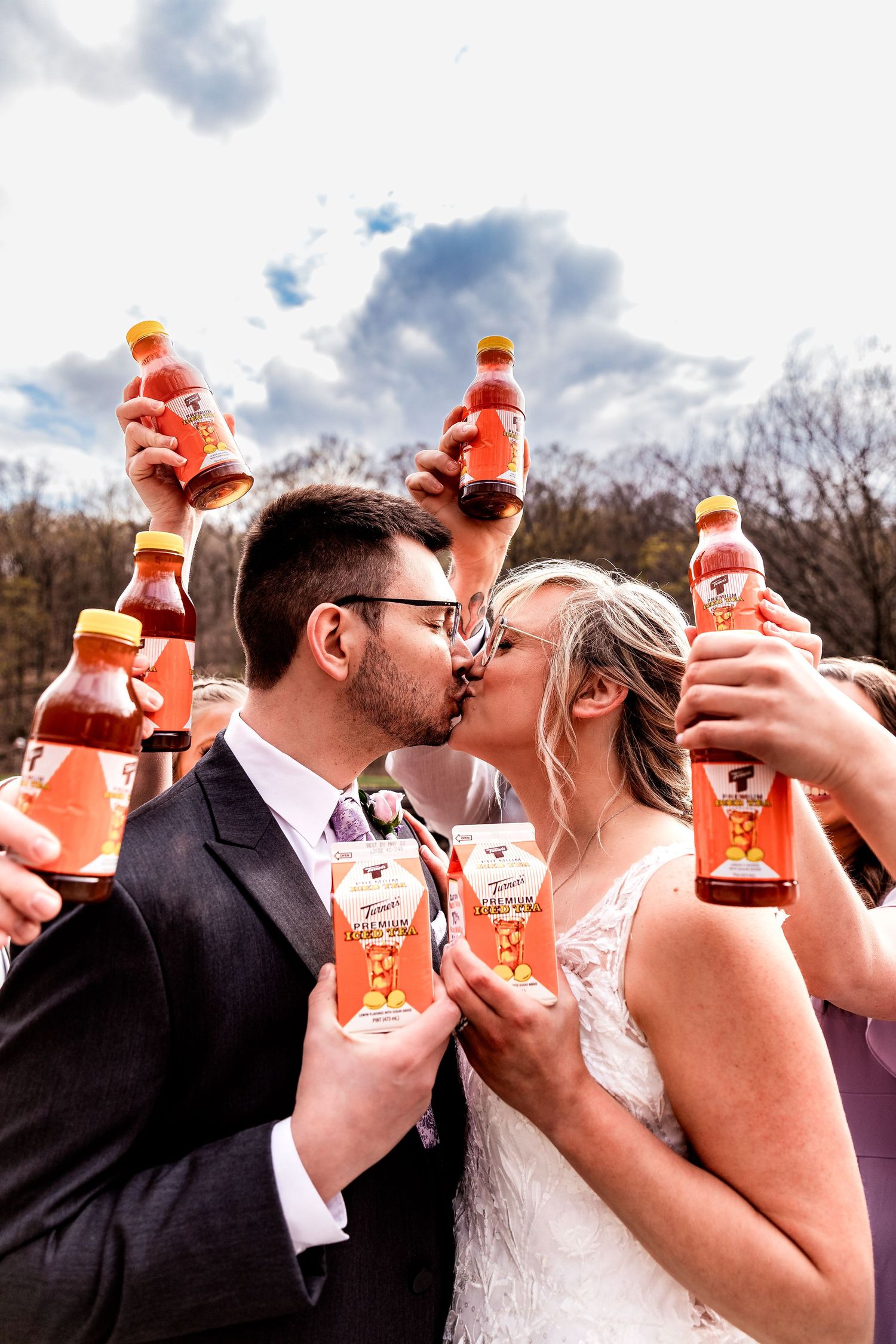 A newlywed couple shares a kiss while wedding guests hold up orange Aperol Spritz bottles against a cloudy sky.