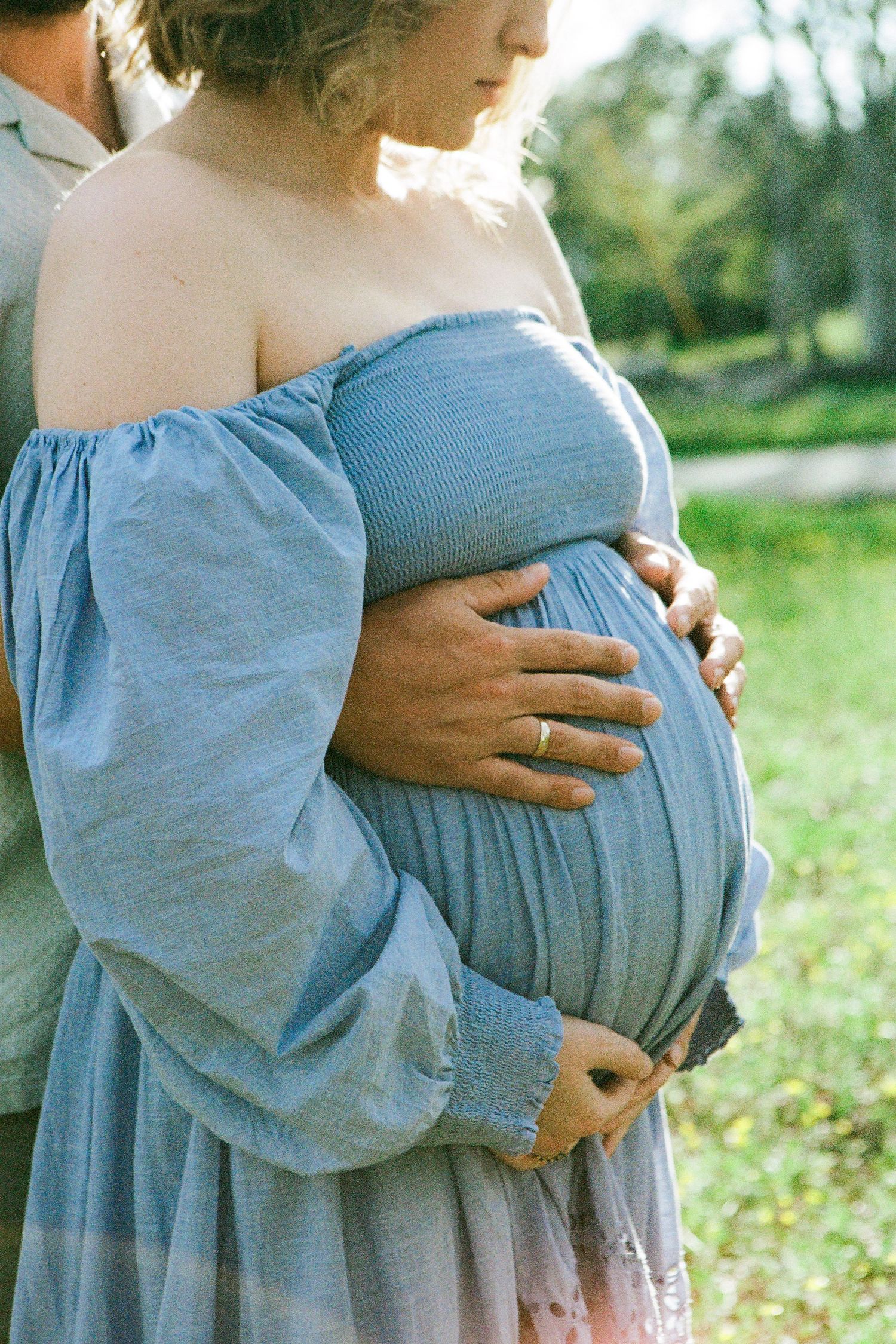 Side view of a pregnant woman in a flowing blue off-shoulder dress outdoors.