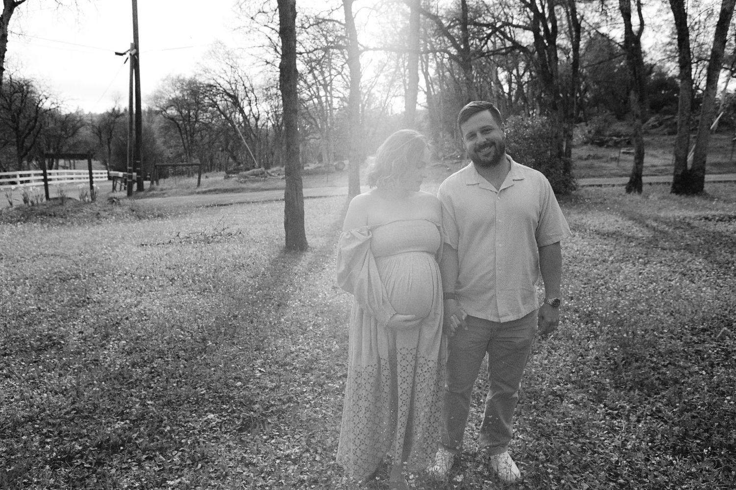 Couple standing together in a sunlit park during pregnancy photoshoot.