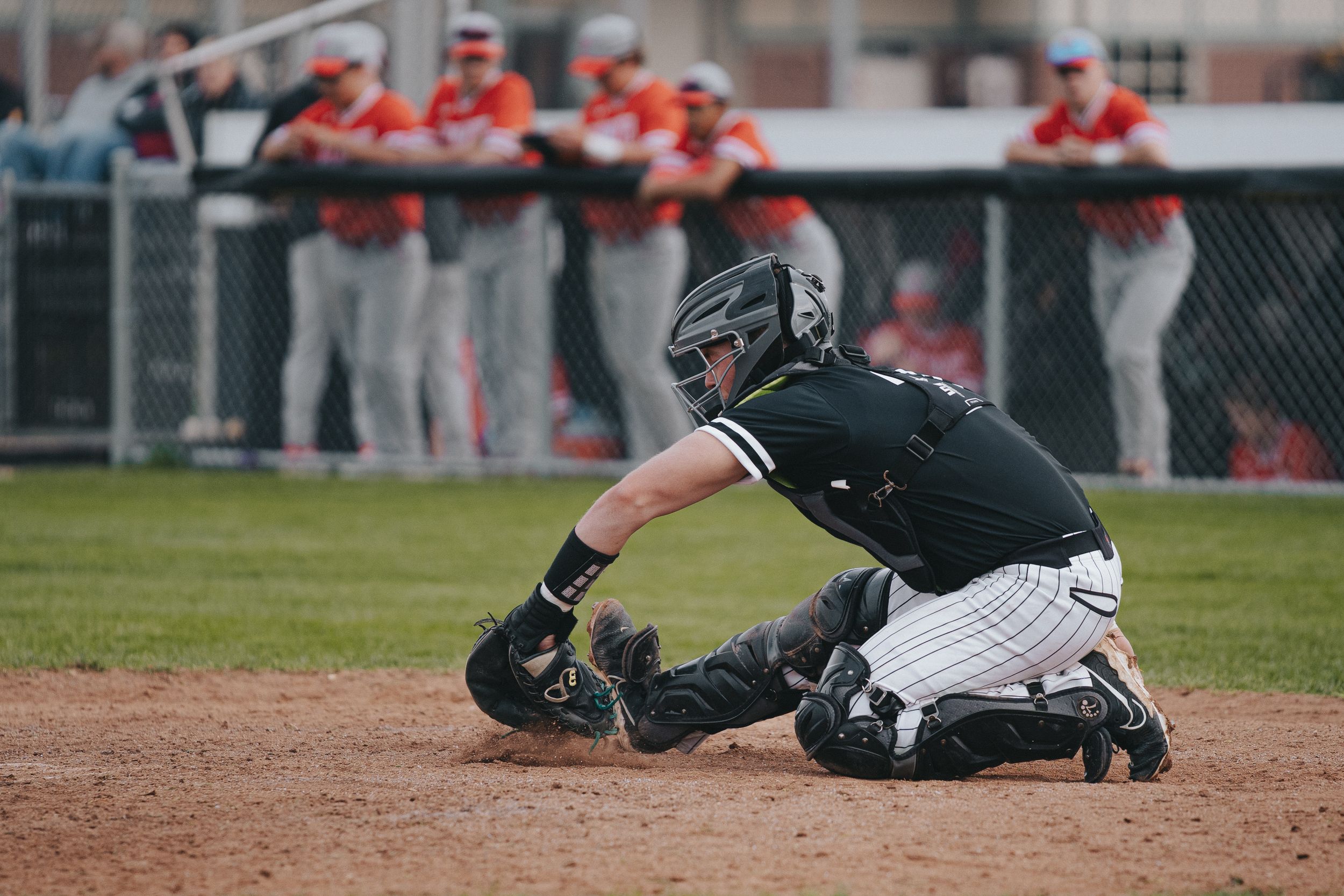 Sports Photography of a a baseball catcher in black uniform reaches low to field a ball during a game - Warsaw, Indiana