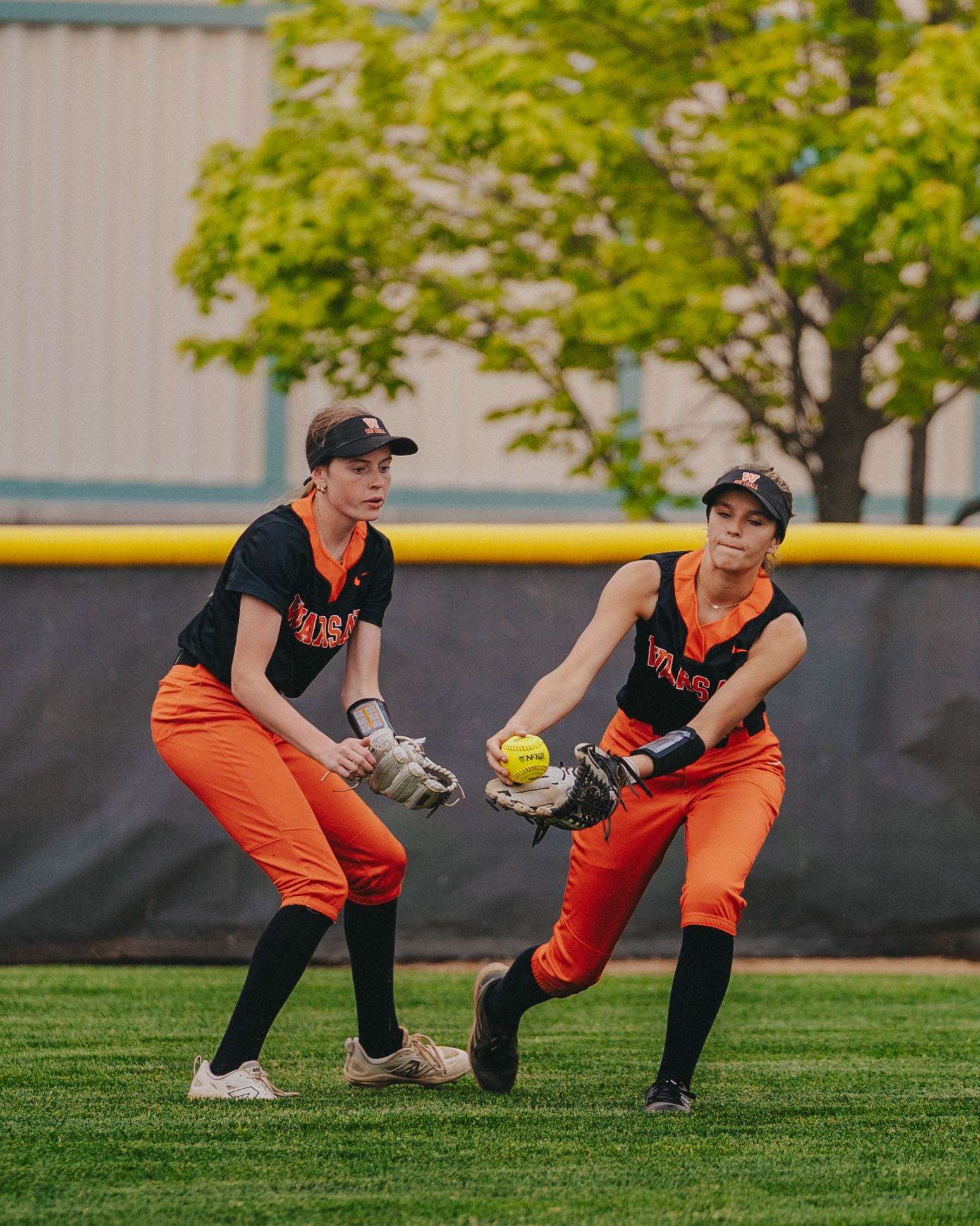 Sports Photography of two softball players in orange uniforms fielding ball during a game on grass field - Warsaw, Indiana