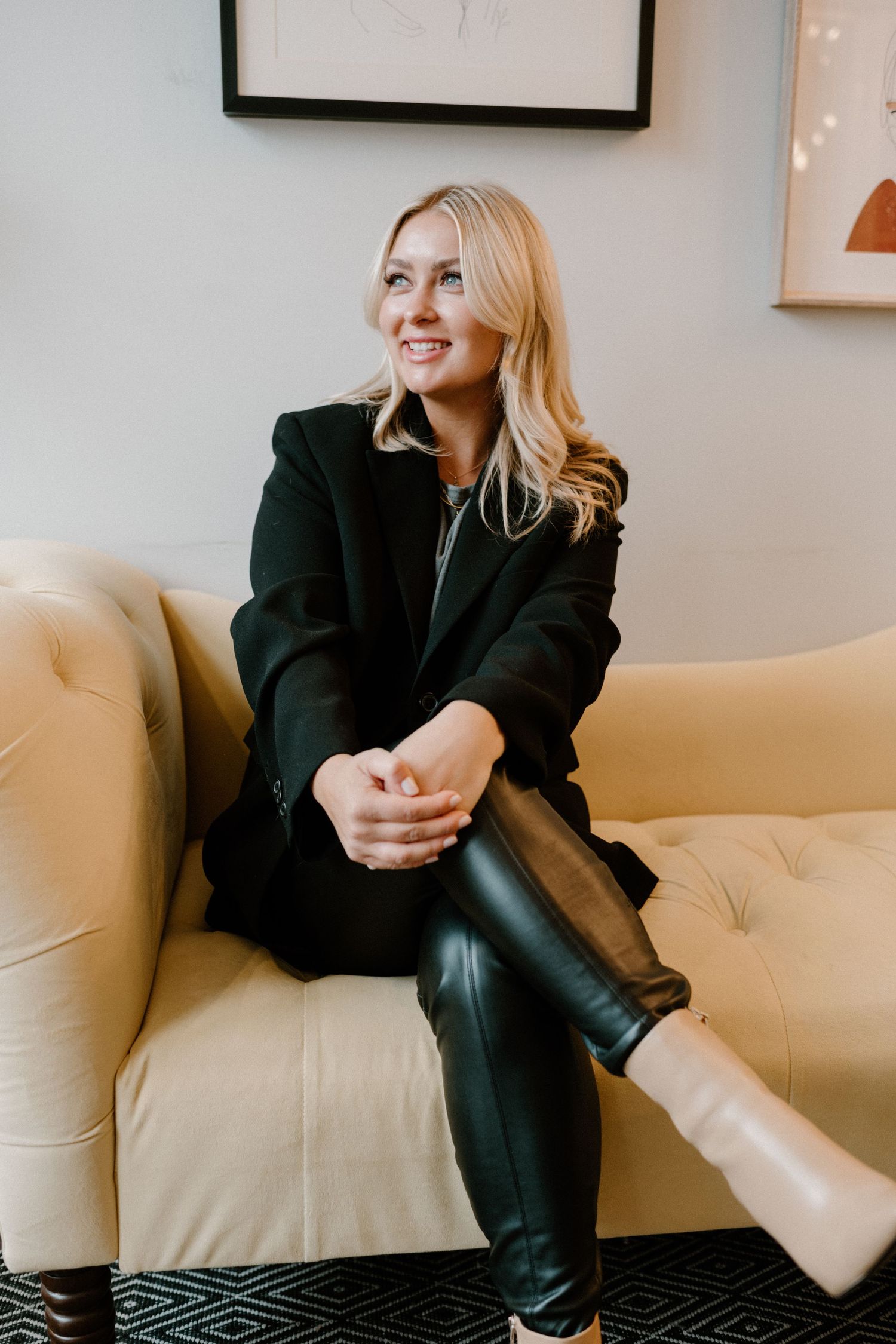Smiling blonde woman in olive athletic wear sits on stool holding dumbbell against brick wall.