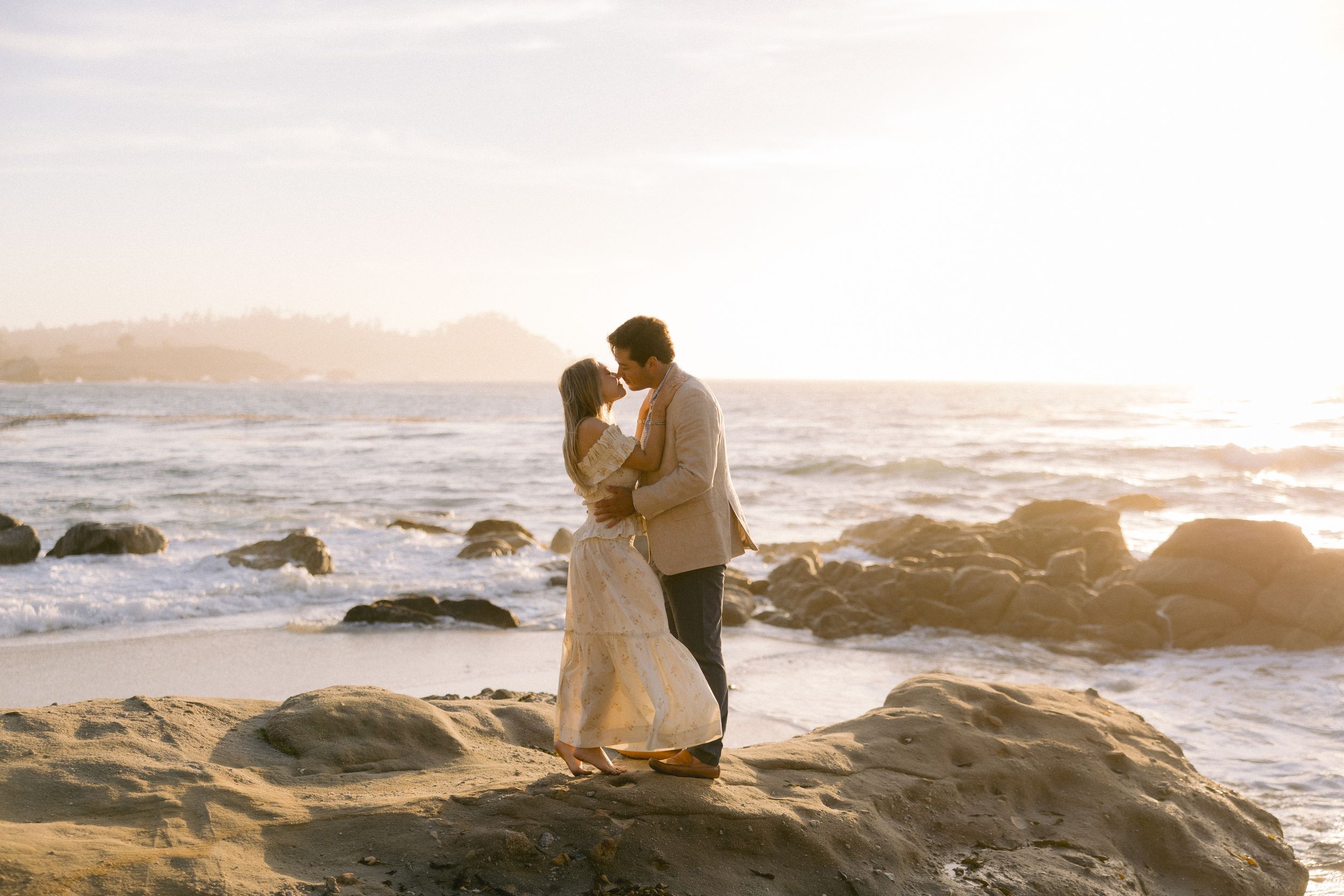 A couple shares an intimate moment at sunset on a rocky beach shoreline with ocean waves crashing nearby.