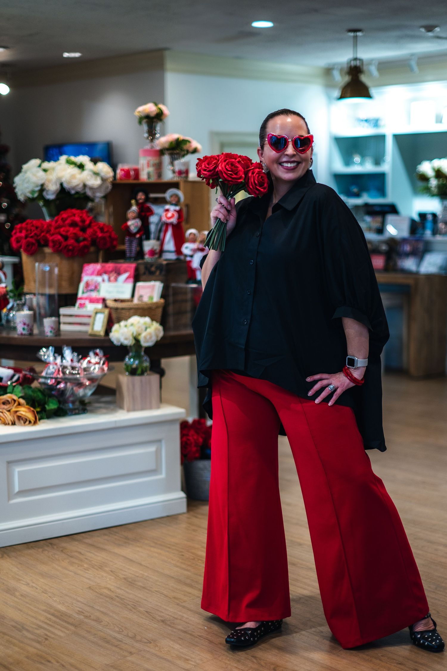Someone wearing a black cape, red wide-leg pants and sunglasses poses in a shop with floral arrangements.