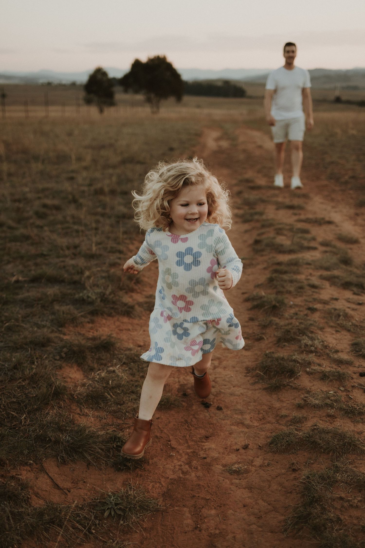 Child in floral dress running joyfully on dirt path through rural field at sunset while her father watches in background.