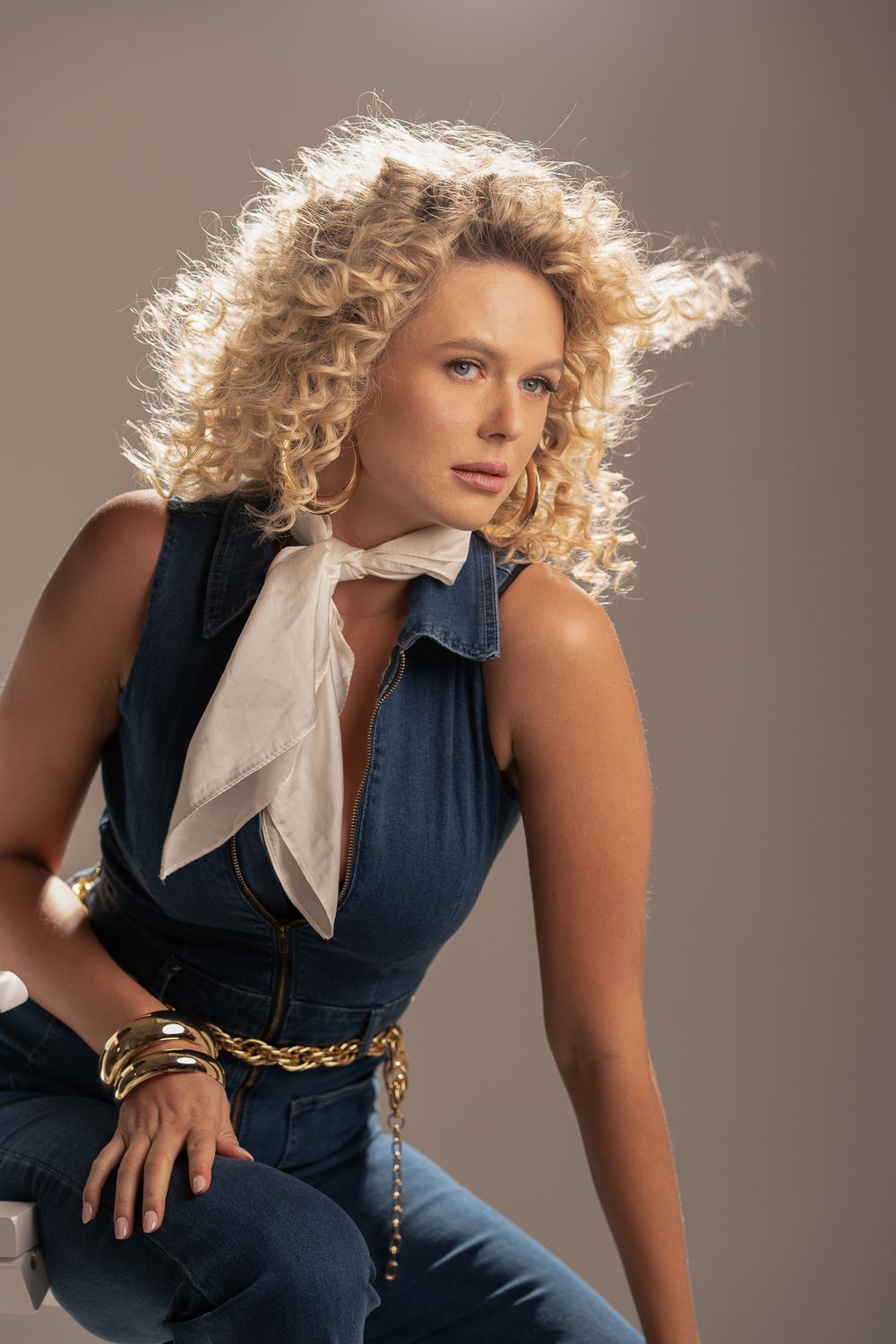 Fashion model wearing denim outfit with white bow tie and curly blonde hair in studio portrait against brown background.